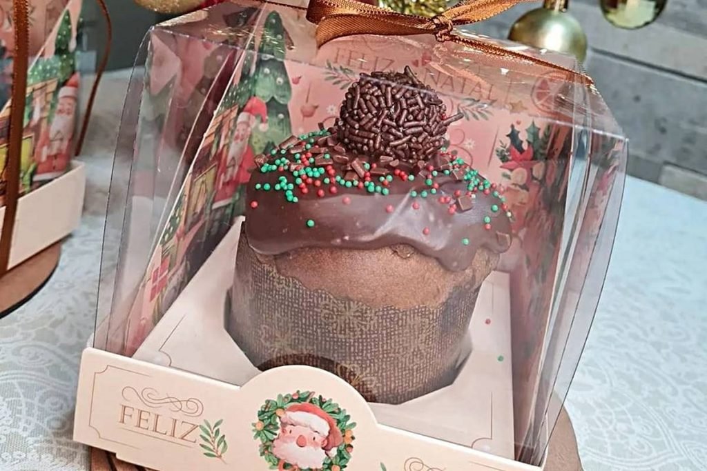 Na foto temos uma caixa com tema de Natal e tampa transparente e dentro um mini chocolate com cobertura marrom escura e granulados verdes e vermelhos e um brigadeiro marrom no centro