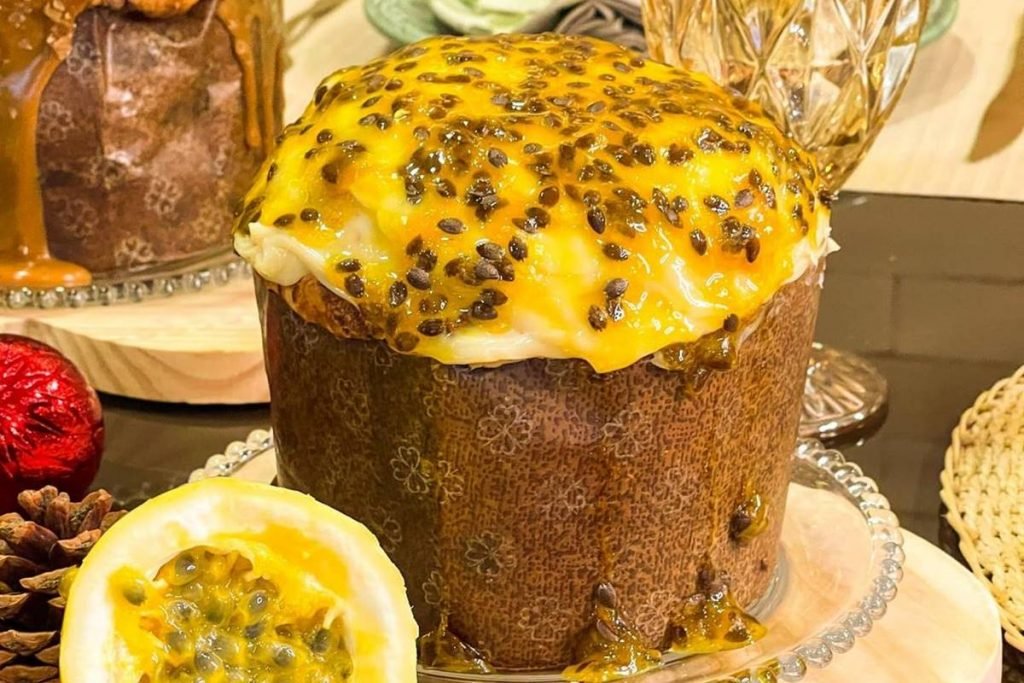 Na foto temos um chocotone coberto com uma calda amarela com pontinhos pretos, que é de maracujá, e uma decoração de natal ao fundo
