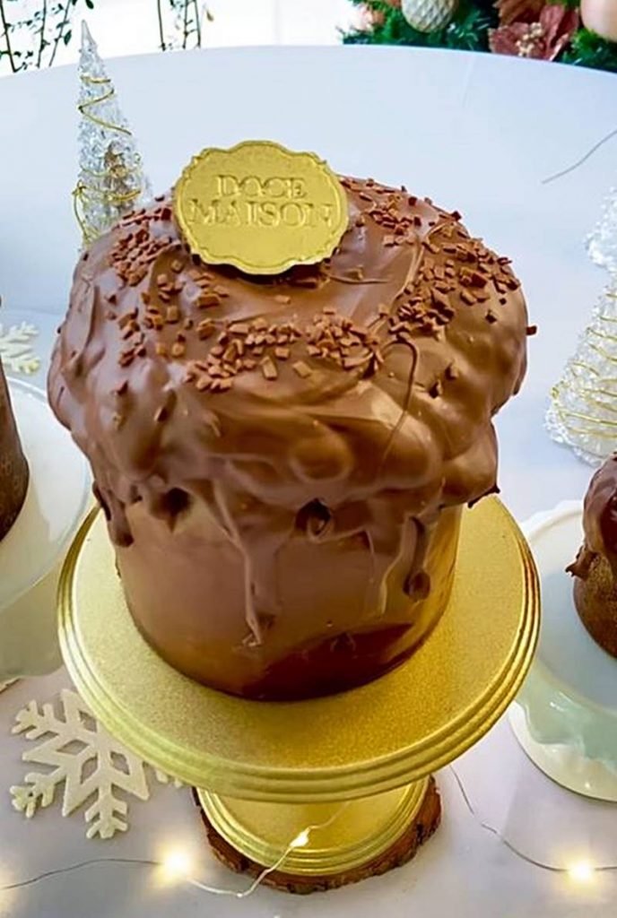 Na foto temos um chocotone coberto por chocolate com um detalhe dourado escrito doce maison em cima sobre uma bailarina dourada