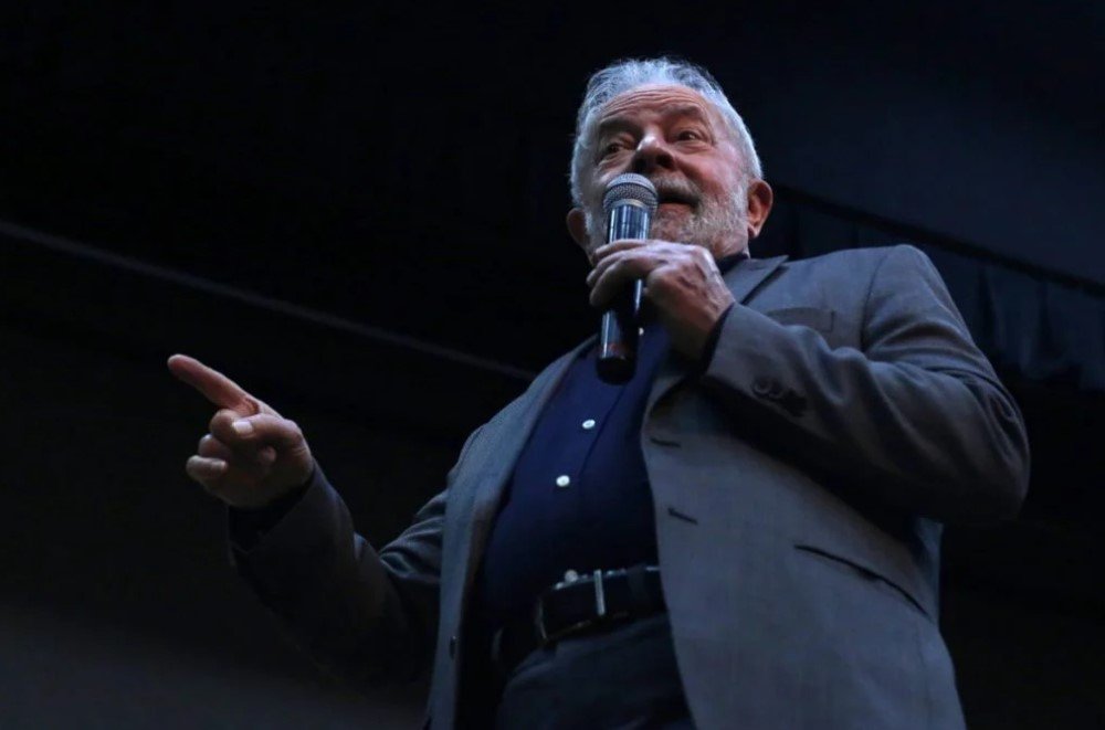 Una foto en color que muestra al expresidente Lula hablando por un micrófono, con el dedo índice de su mano derecha levantado.  Lleva ropa formal y barba.