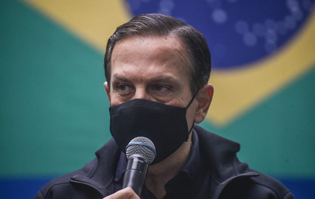 Fotografia colorida de João Doria, governador do estado de São Paulo.  Ele aparece dos ombros para cima, usando máscara preta no rosto.  É um homem branco, de cabelos castanhos penteados para o lado.  Veste moletom azul-escuro e microfone seguro com a mão direita.  Ao fundo, desfocado, a bandeira do Brasil