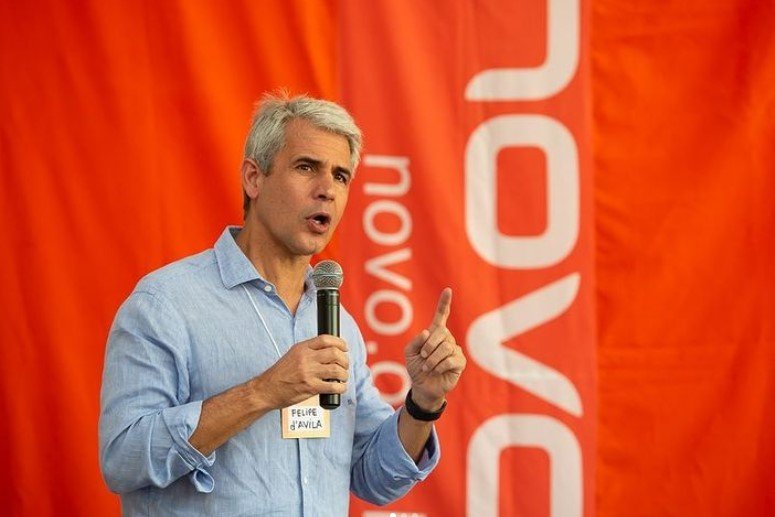 Fotografía en color por Felipe Dávila.  Es un hombre blanco con cabello gris liso en el costado.  En la foto, parece estar hablando por un micrófono, con el dedo índice de su mano derecha levantado.  Lleva una camisa azul claro.  El fondo borroso es naranja y se lee nuevo