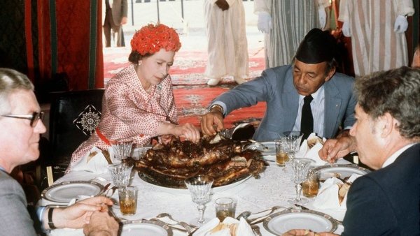 Na fotografia colorida, a rainha Elizabeth II aparece jantando em uma viagem internacional com outras 4 pessoas na mesa