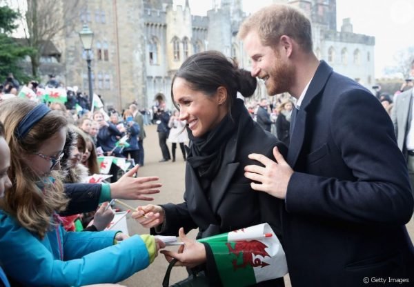 Na fotografia colorida, Meghan Markle e o príncipe Harry aparecem dando autógrafos para os fãs