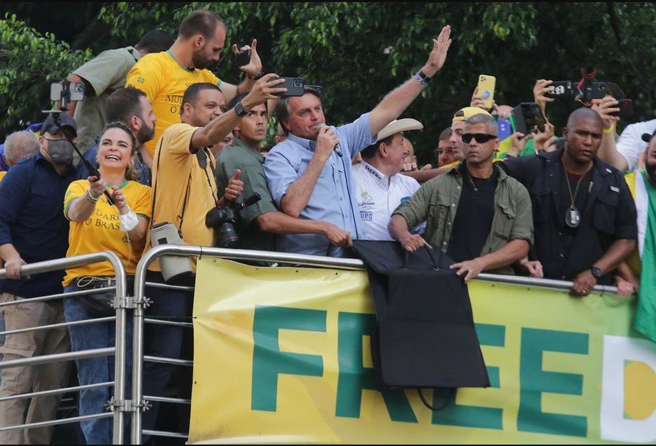 Na imagem colorida, há várias pessoas em cima de um palanque.  Eles usam roupas com a cor da bandeira do Brasil