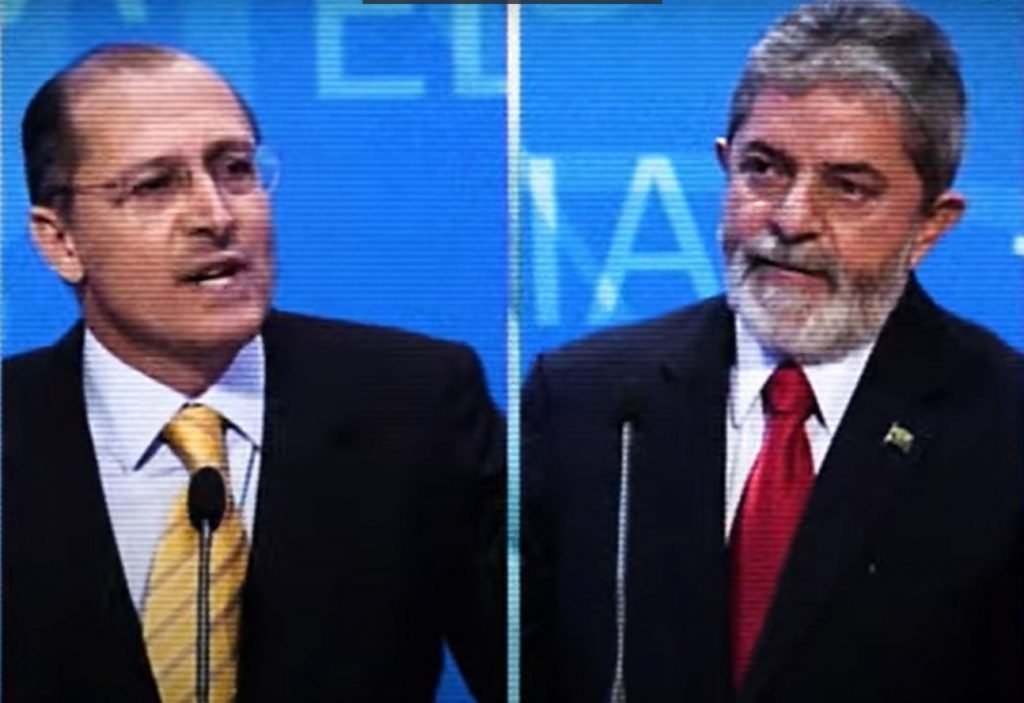 “Lula e Alckmin no debate eleitoral de 2006, aparecem em tela fortaleza enquanto falam.  Ambos cabelo grisalho e usam terno - Metrópoles