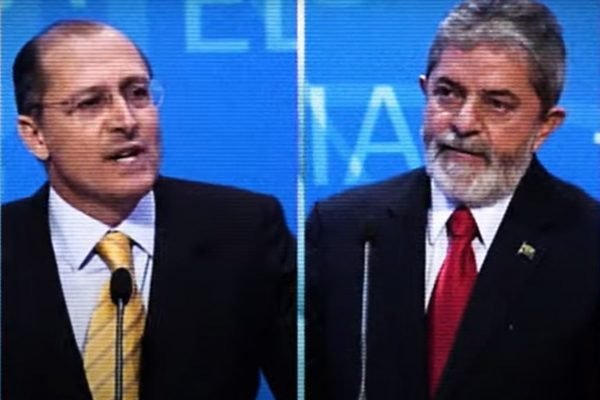“Lula e Alckmin em debate eleitoral de 2006, aparecem em tela dividida enquanto falam. Ambos têm cabelo grisalho e usam terno - Metrópoles