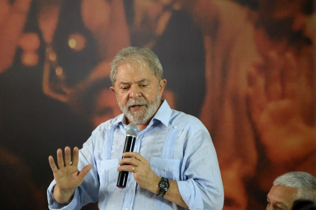 Lula fala ao público com microfone na mão, fazendo gesto com a mão. Ele olha para baixo e usa camisa social sob fundo vermelho - Metrópoles