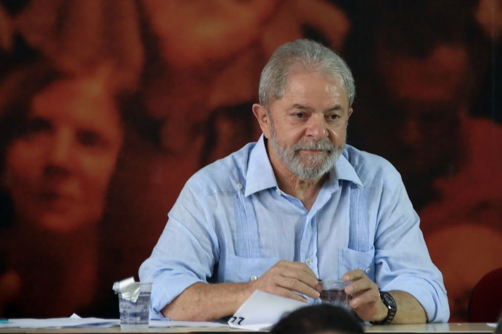 Em evento, Lula está sentado numa mesa e olha para a frente sob fundo vermelho - Metrópoles