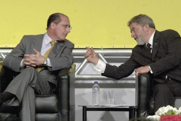 Na foto, sob um fundo amarelo, o então governador de São Paulo, Geraldo Alckmin (PSDB) e o então presidente Lula (PT) conversam sentados em evento - Metrópoles