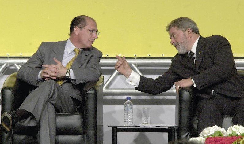 Na foto, sob um fundo amarelo, o então governador de São Paulo, Geraldo Alckmin (PSDB) e o então presidente Lula (PT) conversam sentados em evento - Metrópoles