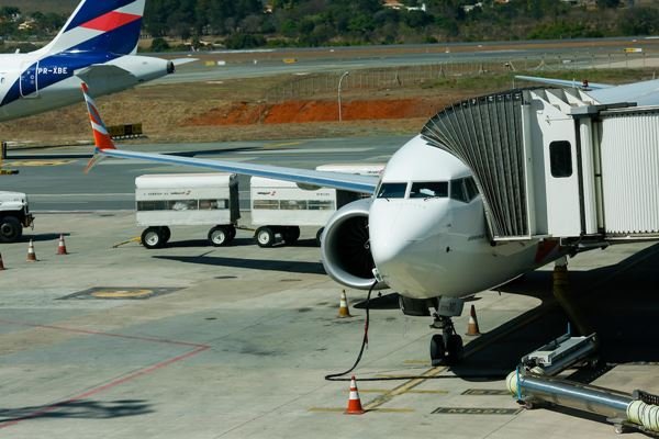 Na imagem colorida, um avião está posicionado à direita. Ele é branco. Atrás há um carrinho de aeroporto