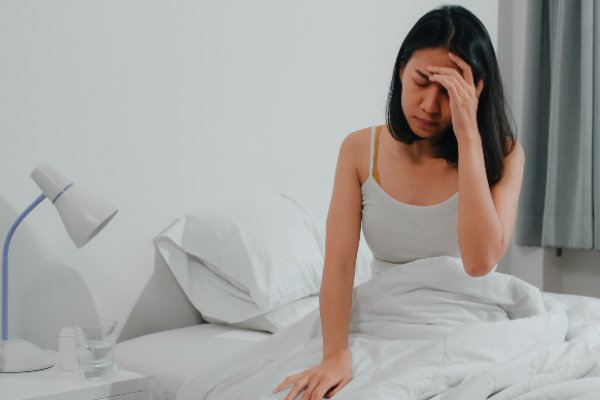 Foto colorida. Mulher oriental, de cabelos pretos e lisos sentada na cama com a mão na cabeça como se tivesse esquecido algo - Metrópoles