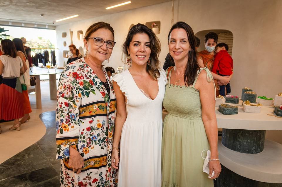 Três mulheres juntas. Uma delas com roupa estampada e as outras duas com vestidos no tom branco e verde 