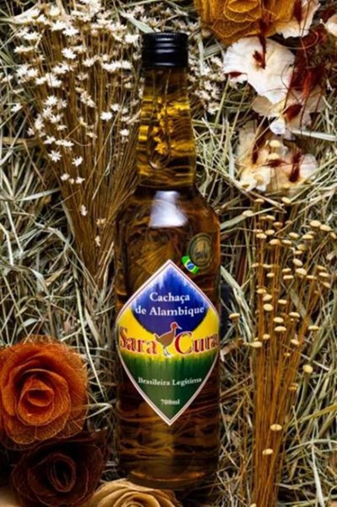 Na foto temos uma cama de flores secas e artesanais e no meio dela uma garrafa com um liquido marrom claro e um rotulo losangular com as cores azul, amarelo e verde escrito sara cura