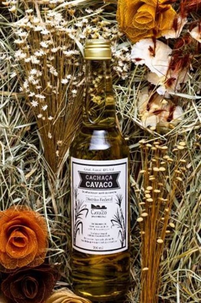 Na foto temos uma cama de flores secas e artesanais e no meio dela uma garrafa com um liquido marrom claro e um rotulo branco com escritas em preto