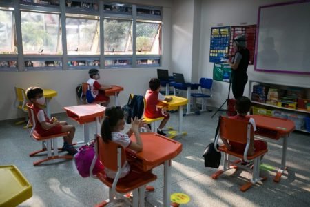 Crianças sentadas em mesas dentro de sala de aula e mulher em frente ao quadro