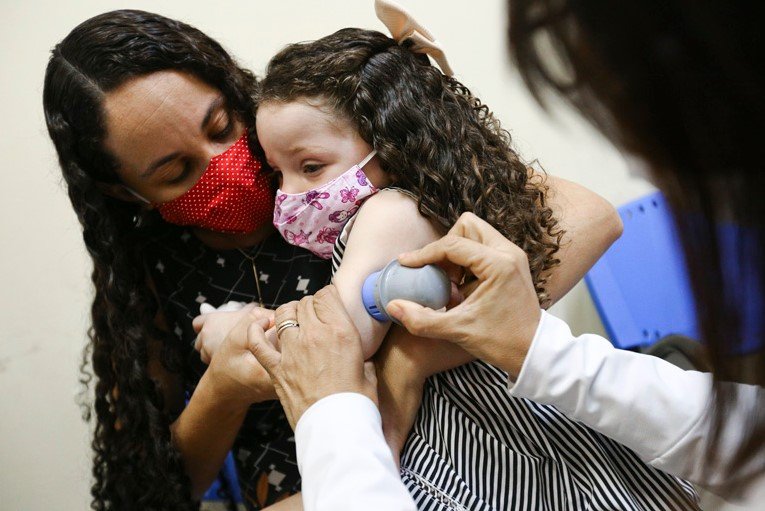 Na imagem colorida, uma mulher com cabelos pretos segura uma criança com cabelos enrolados no colo. Alguém aplica um aparelho para diabetes no braço da criança