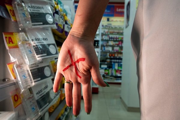Na fotografia colorida, a mulher mostra um x vermelho na mão em uma farmácia