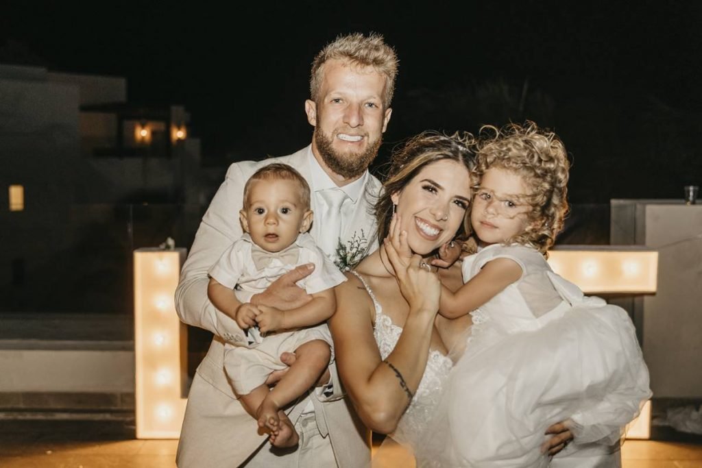 dois adultos brancos vestidos de branco com duas crianças, um no colo do homem e outra no colo da mulher. todos sorrindo e posando para um retrato