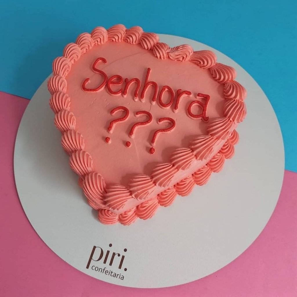 Na foto temos um bolo em forma de coração na cor rosa com a palavra senhora e três pontos de interrogação escritos de vermelho