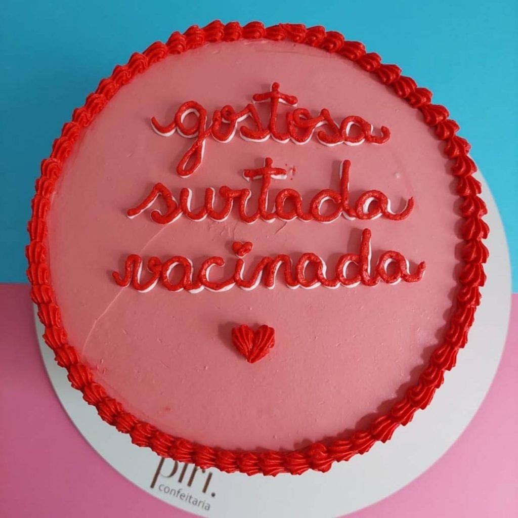 Na foto temos um bolo redondo rosa com as palavras gostosa, surtada e vacinada escritas em vermelho