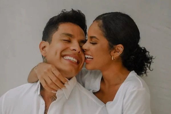 Fotografia colorida. Victor e Sthe posam abraçados de chamego. Ele sorri e ela finge dar uma mordida em sua bochecha