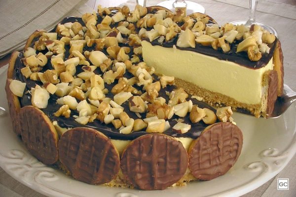 Na foto temos um prato branco redondo com uma torta com uma camada de biscoitos, outa de creme branco e cobertura de calda marrom escura de chocolate e castanhas picados e ao redor dela, biscoitos cobertos de chocolate