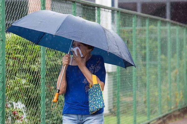 Fotografia colorida. Mulher anda na chuva de guarda-chuva e mão na boca com pano