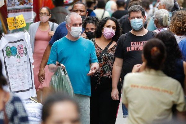 Fotografia colorida. Multidão aparece andando na rua com máscara