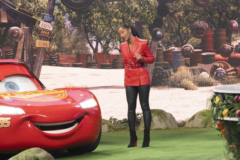 Na foto temos uma mulher negra jovem com um vestido vermelho de manga longa, meias pretas e botas pretas olhando para uma replica do relampago mcqueen de carros