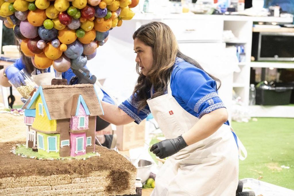 na foto temos uma mulher branca de camiseta azul, avental branco e luvas pretas mexendo em uma casinha com diversas bolas coloridas em cima simulando baloes