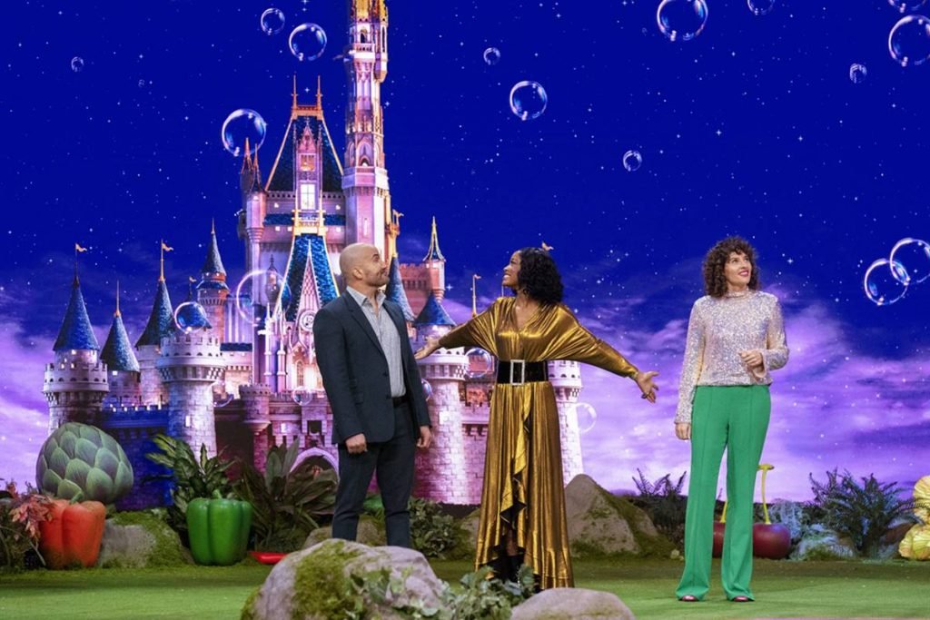 Na foto temos ao fundo o castelo da cinderela projetado e na frente tres pessoas: um homem branco careca de terno cinza escuro, uma mulher negra de vestido dourado e uma mulher branca com blusa branca e calça verde