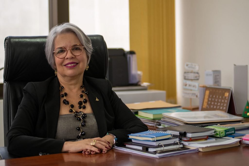 Mulher de óculos e cabelos grisalhos sentada em cadeira preta atrás de mesa com livros