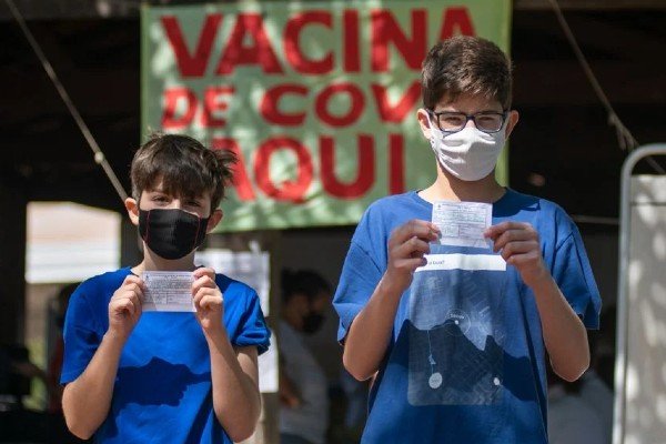 fotografia colorida.  Jovens em camisas azuis seguram registros de vacinação em frente à placa do centro de vacinação