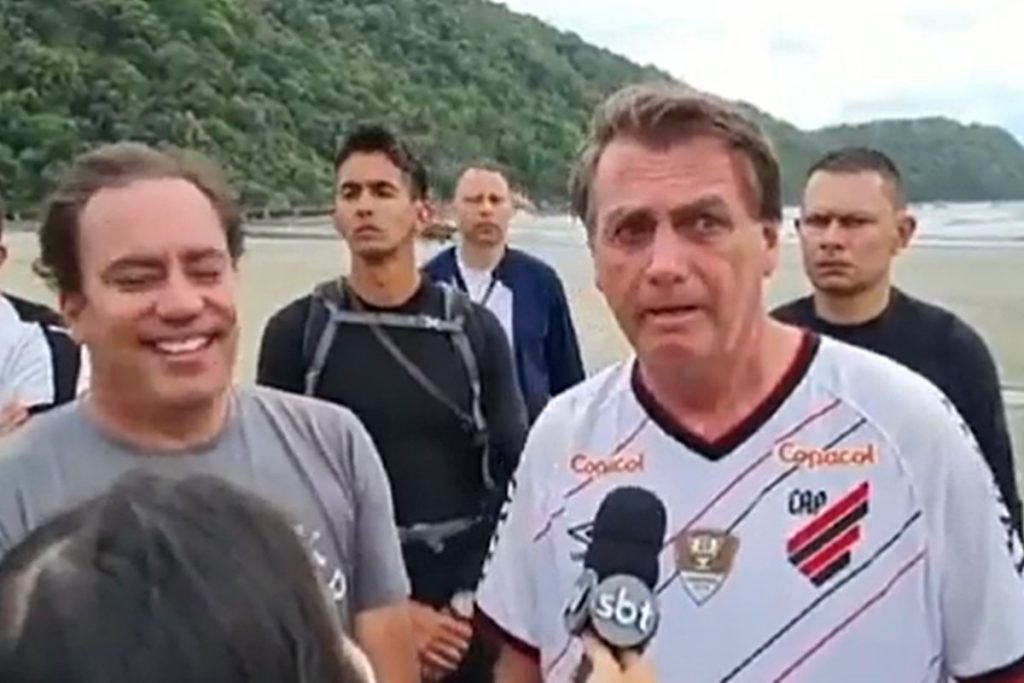 Na foto temos ao fundo uma praia, e na frente o presidente jair bolsonaro e algumas pessoas ao redor