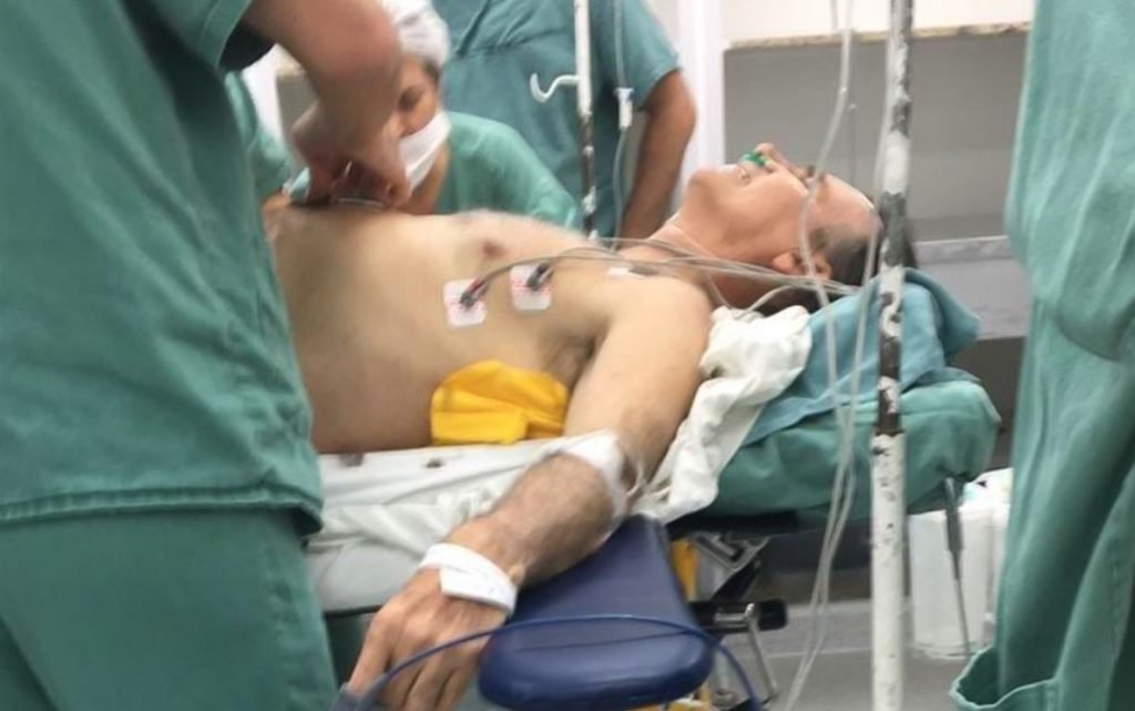Na imagem colorida, um homem está deitado em uma maca de hospital. Ele está sem blusa e há aparelhos ligados a ele. Pessoas com roupas verdes estão ao redor com as mãos sobre a barriga do homem
