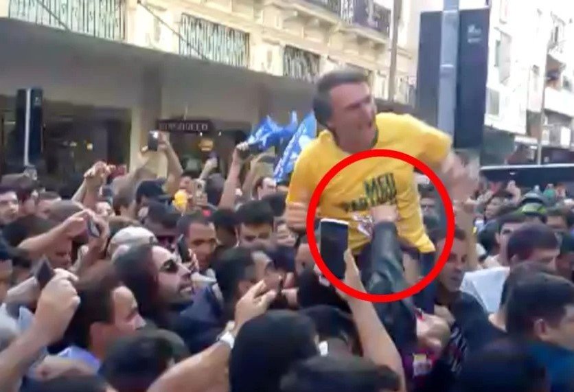 Na imagem colorida, uma multidão segura um homem com blusa amarela. O homem faz uma cara de dor e um circulo vermelho mostra uma mão atingindo a barriga do homem