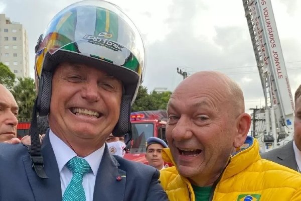 Fotografia colorida. Luciano e jair posam para fotografia sorridentes em meio à multidão. Bolsonaro está com um capacete e Hang com uma jaqueta do Brasil
