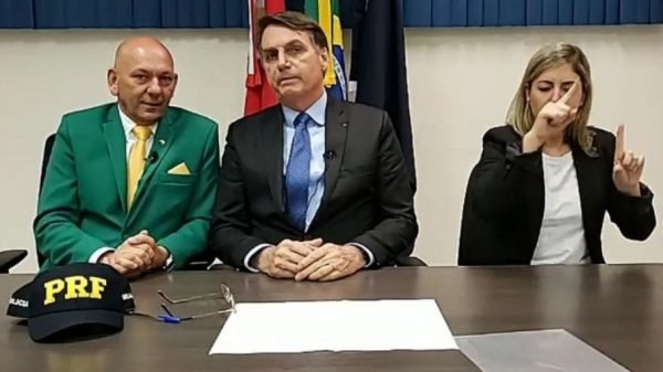 Fotografia colorida. Luciano e Bolsonaro aparecem sentados lado a lado em live. Do lado direito da imagem, uma mulher se senta junto aos dois homens
