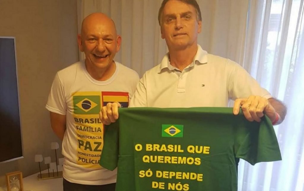 Fotografia colorida.  Bolsonaro e Hang posam para foto.  Jair segura camiseta com as cores da bandeira do Brasil