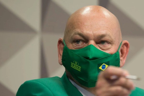 Fotografia colorida. Luciano depõe na CPI vestido com paletó verde e máscara com a bandeira do Brasil. Na imagem ele aponta o dedo para a frente