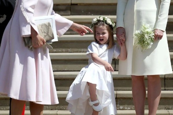 Fotografia colorida. Charlotte posa de mãos dadas com a mãe no casamento de Meghan