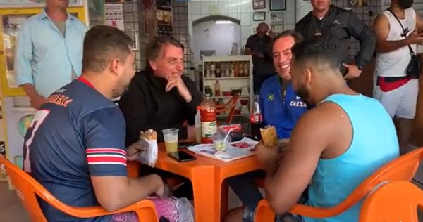 Bolsonaro sentado em uma mesa de plástico com o presidente da Caixa e dois apoiadores