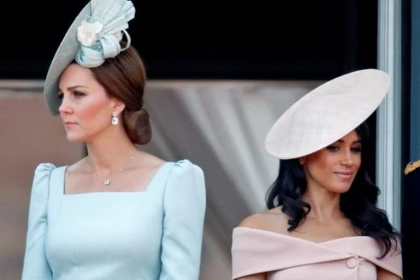 Fotografia colorida. Meghan e Kate aparecem lado a lado. Uma com o rosto virado pra cada lado