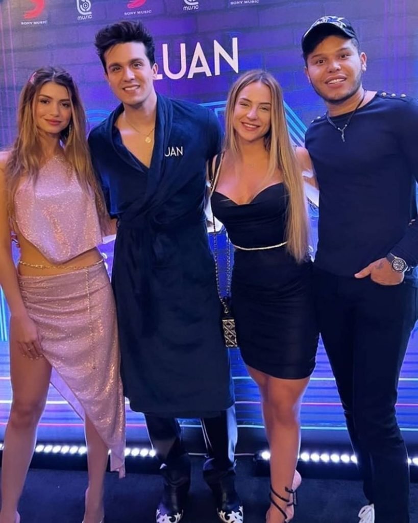 Na foto temos uma mulher branca de cabelo longo com roupa brilhosa, um homem branco de roupão azul, uma mulher loira e branca de vestido preto e um homem de pele morena de camiseta azul manga longa, calça preta e boné