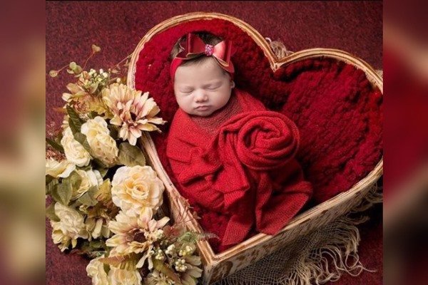 Na imagem, um bebê está posicionado dentro de um cesto colorido em formato de coração.  Ele está embalado por manta vermelha e há flores ao lado esquerdo do cesto