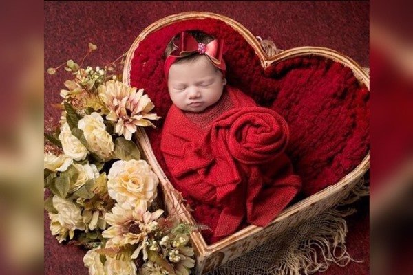 Na imagem colorida, um bebê está posicionado dentro de um cesto em formato de coração. Ele está embalado por manta vermelha e há flores ao lado esquerdo do cesto