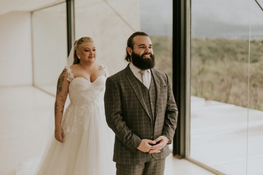 Pastel com caldo de cana: casal comemora união em pastelaria no DF - Veja fotos 6 Mulher vestida de noiva e homem vestido de noivo com braços dados em frente a uma janela de vidro