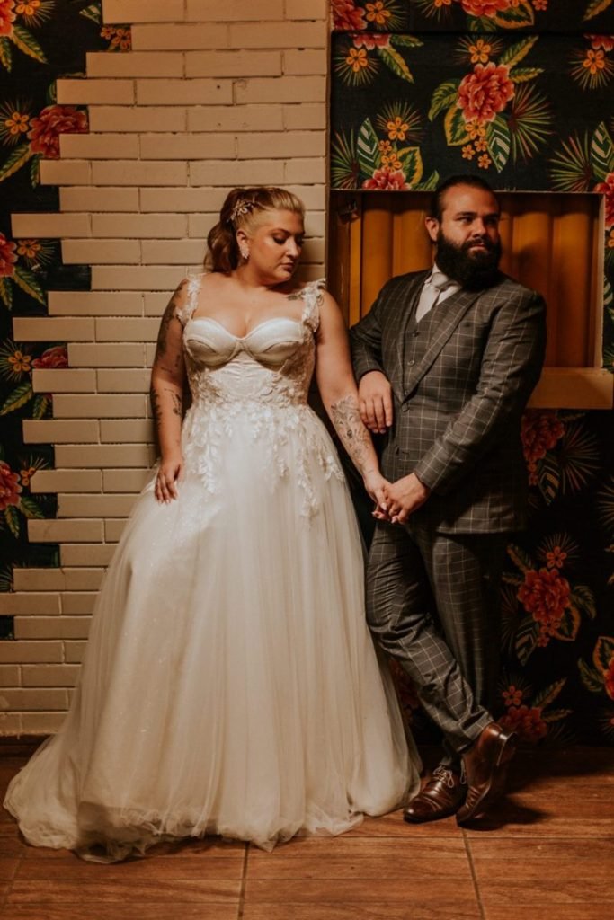 Pastel com caldo de cana: casal comemora união em pastelaria no DF - Veja fotos 3 Vestidos de noiva e noivo, casal se abraça encostados a uma parede de tijolos